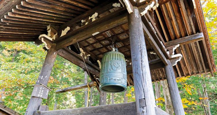 Campana tradicional de templo japonés colgando bajo un techo de madera con follaje otoñal de fondo