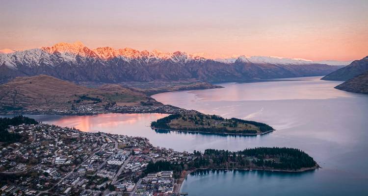 Vue aérienne à l'aube sur Queenstown, le lac Wakatipu et les Remarkables aux sommets enneigés rougeoyant de rose à la lumière matinale.