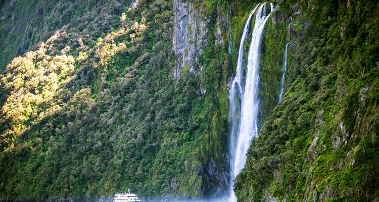 Une haute cascade se précipite le long des falaises verdoyantes de Milford Sound avec un petit bateau de croisière qui paraît minuscule en dessous.