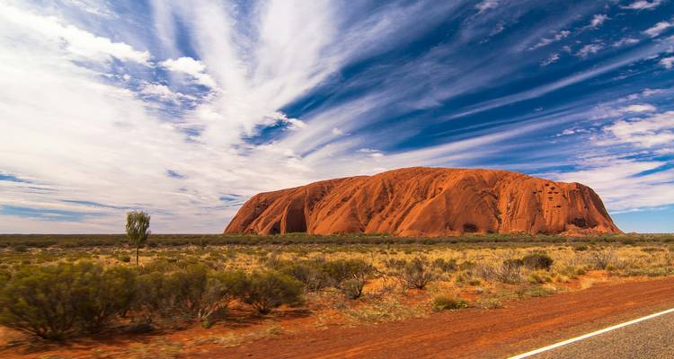 Le monolithe massif de grès rouge d'Uluru s'élève au-dessus d'une brousse aride sous des nuages spectaculaires aux trainées dramatiques.