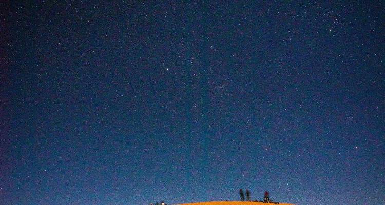 Sternenerfüllter Nachthimmel über einer Wüstendüne mit kleinen silhouettierten Gestalten, die auf dem Kamm sitzen.
