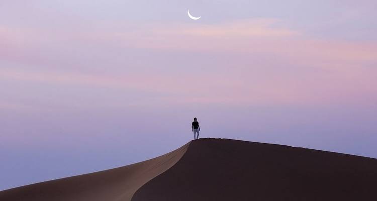 Silhouet van eenzame persoon op duinkam bij schemering met sikkelmaantje in pastelkleurige lucht