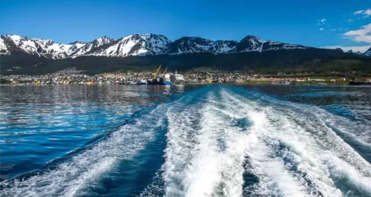 Le sillage d'un bateau s'étend vers Ushuaia avec des pics couverts de neige se reflétant dans la baie.