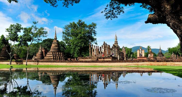 Las antiguas estupas de ladrillo y las agujas de Sukhothai Historical Park reflejadas en un estanque tranquilo con exuberante vegetación y cielos azules.