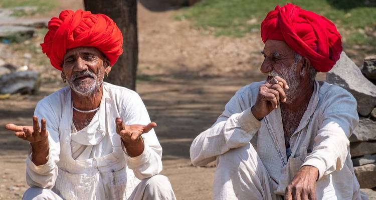 Zwei Männer in traditioneller indischer Kleidung sitzen auf dem Boden und führen ein Gespräch.