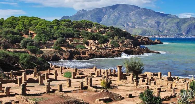 Ancient coastal ruins with stone columns and foundations set against turquoise sea and forested hills in Algeria.