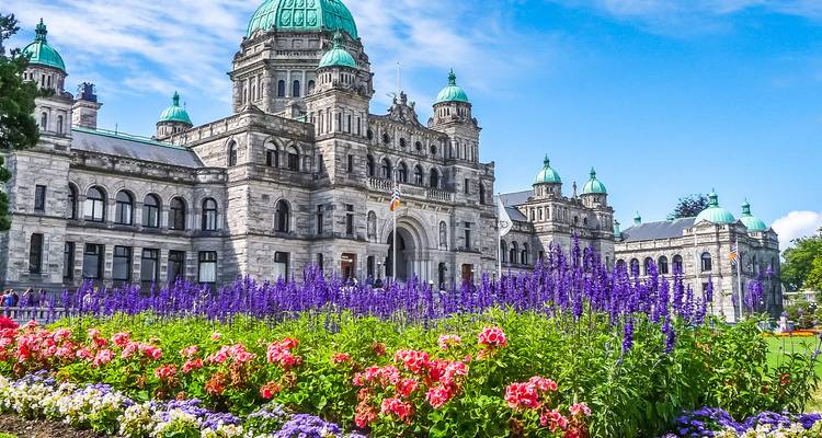 Les bâtiments du parlement victorien avec des dômes turquoise derrière des parterres de fleurs vibrants sous un ciel bleu éclatant.