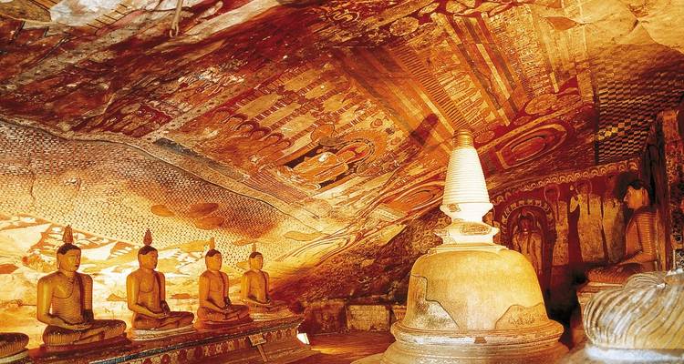 Golden-lit interior of Dambulla cave temple with seated Buddha statues and ornate painted ceiling
