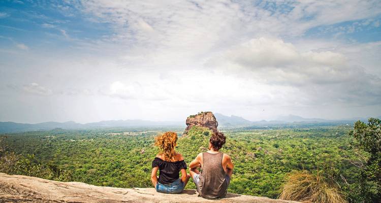 Couple sits on a rocky ledge overlooking the lush plain and iconic Sigiriya Rock fortress.
