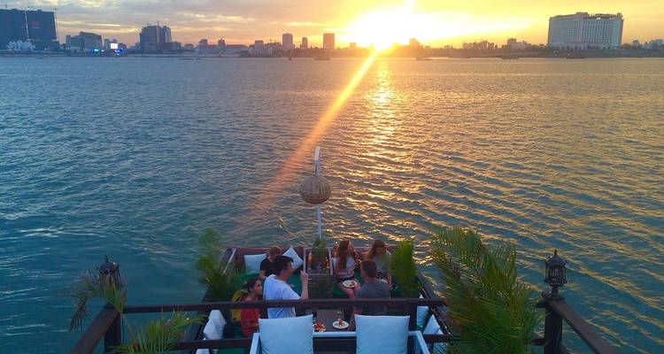 Gruppen von Menschen, die bei Sonnenuntergang auf einem Boot speisen mit Blick auf die Skyline einer Stadt.