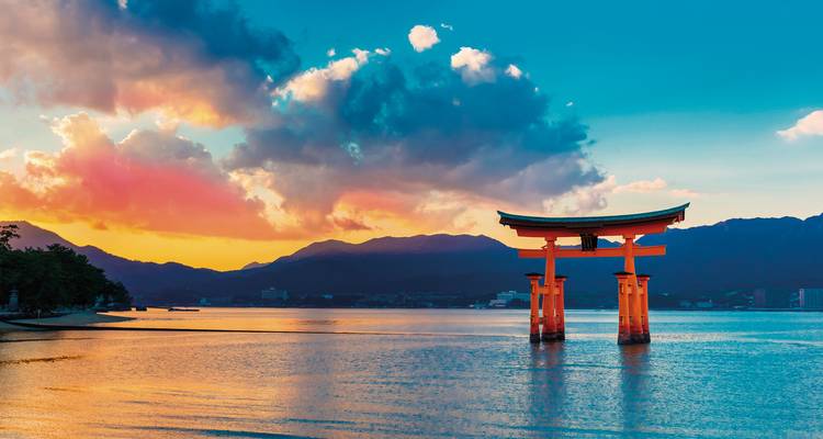 Schwimmendes Torii-Tor bei Sonnenuntergang mit bunten Wolken und Bergen.
