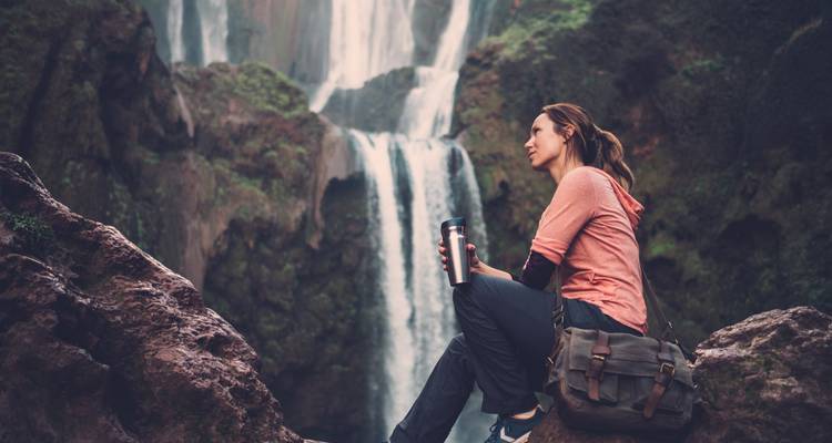 Frau sitzt mit einem Getränk in der Nähe eines Wasserfalls.