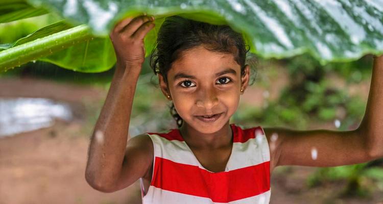 Smiling young girl shelters from rain under a giant tropical leaf, water droplets glistening.