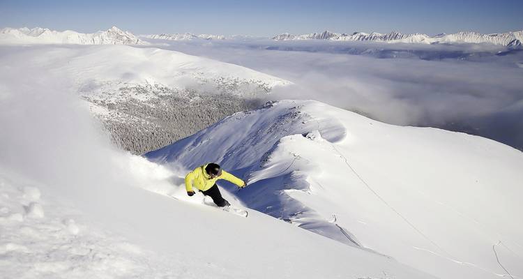 Skier carving through fresh powder on a steep, untouched snowy slope above clouds.