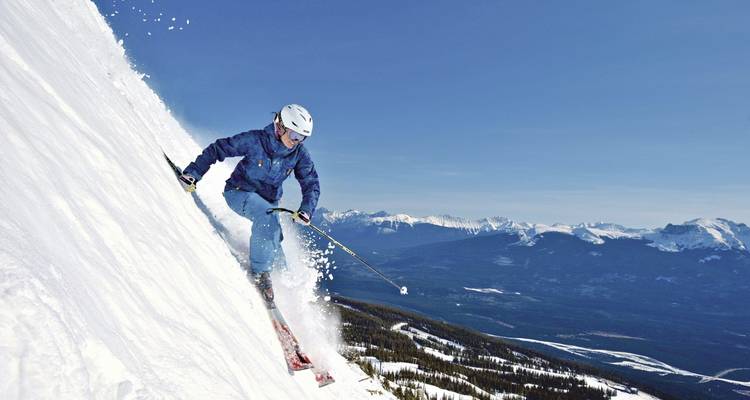 Action shot of skier descending a very steep snowy face with peaks in background.