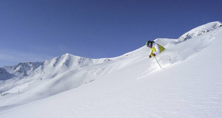 Skier in bright gear making a powder turn on wide open snowy slope beneath blue sky.