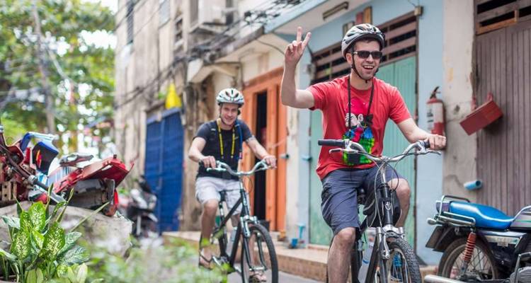 Zwei fröhliche Radfahrer erkunden eine enge Stadtgasse, die mit lebendigen Türen und Rollern gesäumt ist.