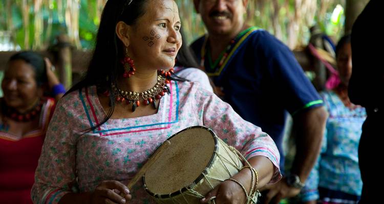 Indigenous woman in patterned dress plays a hand drum during a traditional gathering.