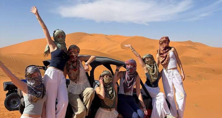 Grupo de mujeres con velos y ropa ligera celebrando en la arena del desierto junto a un vehículo todoterreno.