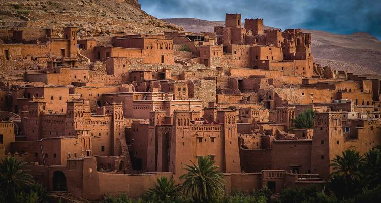 Sonnenbeschienene Lehm-Zitadelle von Ait Benhaddou vor einer felsigen Wüstenhügellandschaft.