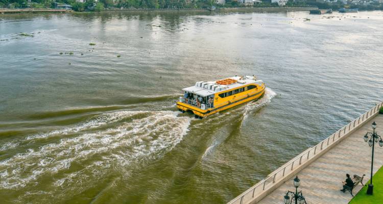 Bateau touristique jaune naviguant sur une rivière avec des bâtiments et de la verdure en arrière-plan.