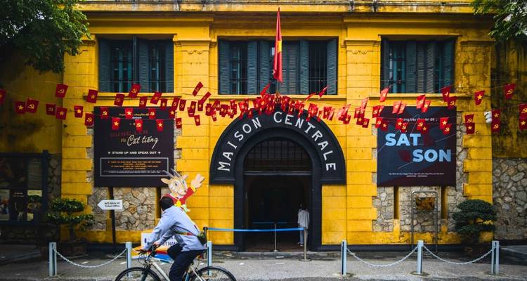 Bâtiment historique avec façade jaune et un vélo qui passe, avec des drapeaux suspendus au-dessus.