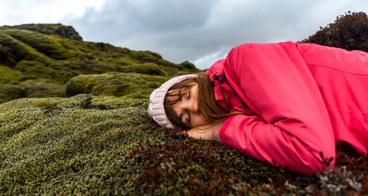 Reisender in rosa Jacke liegt auf schwammigem grünem Moos eines Lavafeldes unter grauem Himmel.