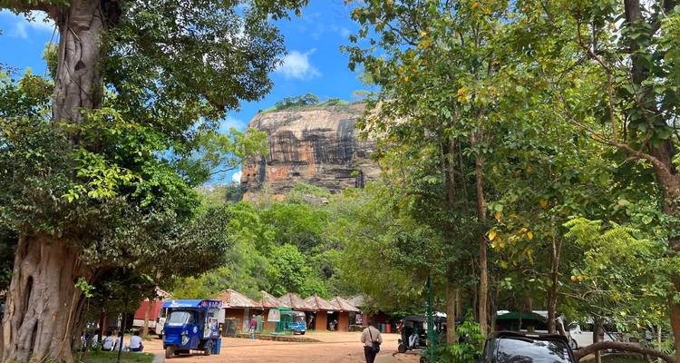 Une forêt dense encadre le monolithe rocheux emblématique de Sigiriya vu depuis une aire de stationnement en terre battue avec des tuk-tuks.