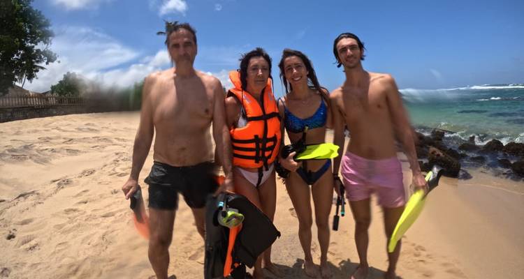 Quatre plongeurs en apnée en tenue de plongée posent sur une plage de sable fin avec des vagues tropicales en arrière-plan.