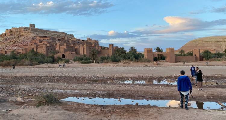 Aït-Ben-Haddou avec des personnes marchant au premier plan, encadré par une rivière et des collines.