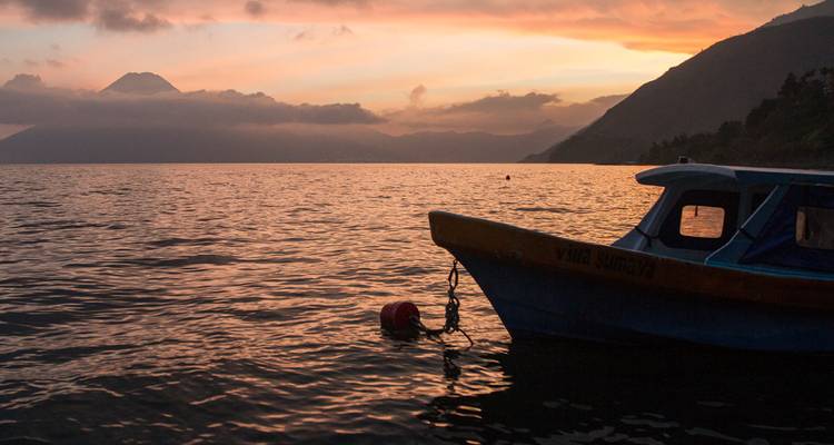 Small boat floats on a tranquil lake as volcano silhouettes fade into a glowing sunset sky