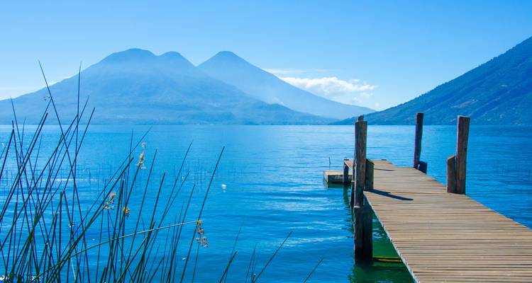 Muelle de madera que se extiende en el brillante lago azul Atitlán con volcanes gemelos a la distancia.