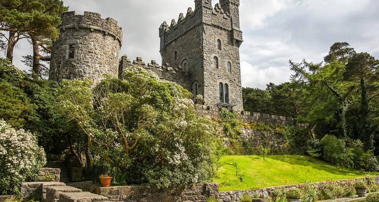 Stone castle tower surrounded by lush gardens and tall pines under an overcast sky in Ireland.
