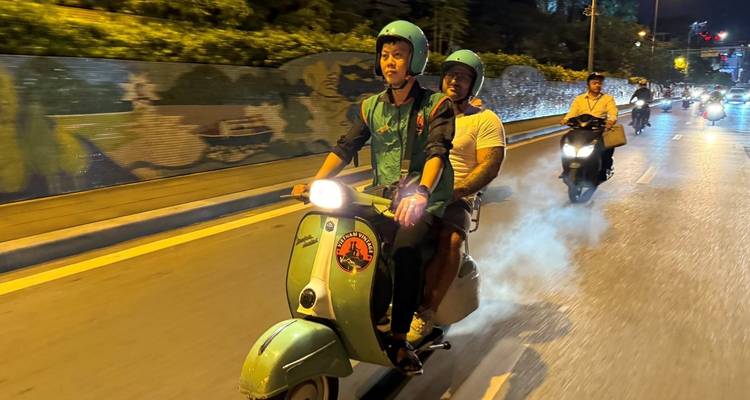 Zwei Personen auf einem Roller fahren nachts auf einer Straße.