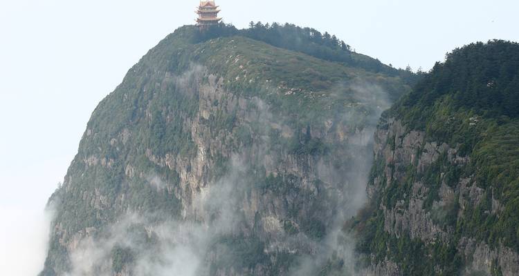 Nebelige Klippen des Mount Emei mit einem kleinen Pagodentempel, der auf dem Gipfel über treibenden Wolken thront.