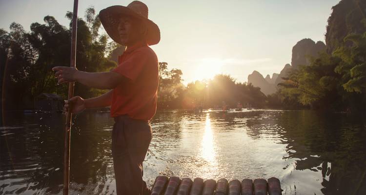 Ein Bambusfloß-Bootsmann stakt mit einer Stange entlang eines ruhigen Flusses bei Sonnenuntergang inmitten der Karstgipfel von Yangshuo.
