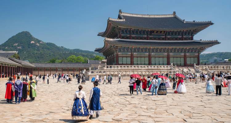 Amplio patio de un palacio real coreano con turistas vistiendo coloridos hanbok y el salón principal bajo un cielo azul despejado.