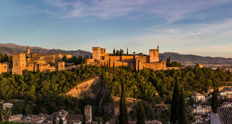 Vista panorámica de la fortaleza de la Alhambra brillando con la luz cálida del atardecer, con las montañas al fondo y las copas de los árboles abajo.