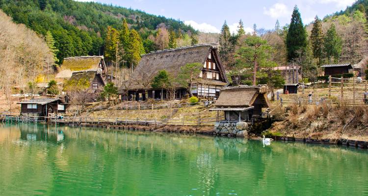 Casas de granja con techo de paja tradicional reflejadas en un lago esmeralda rodeado de colinas boscosas.