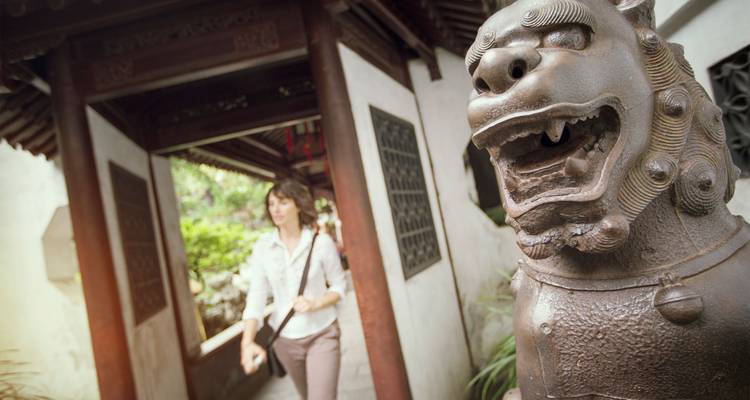 Una mujer pasea junto a una gran estatua de león guardián de bronce en un corredor de jardín clásico chino.