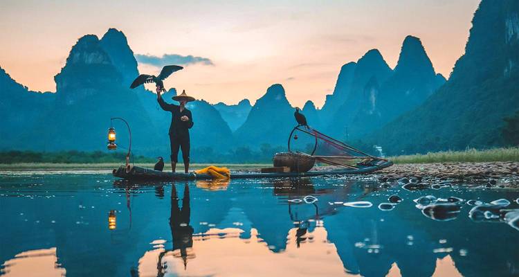 Le pêcheur de cormorans se tient sur un radeau de bambou au crépuscule, entouré d'une eau semblable à un miroir et de pics karstiques.