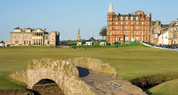 Un histórico puente de piedra para golf en primer plano con los imponentes edificios del club de un famoso campo de links.