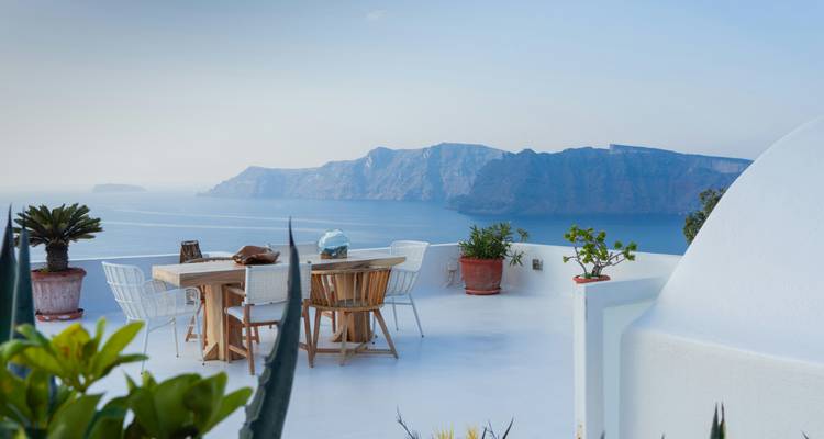 Terrasse minimaliste blanche avec ensemble de salle à manger offrant une vue sur la caldeira de Santorin et les falaises lointaines à la lumière douce.