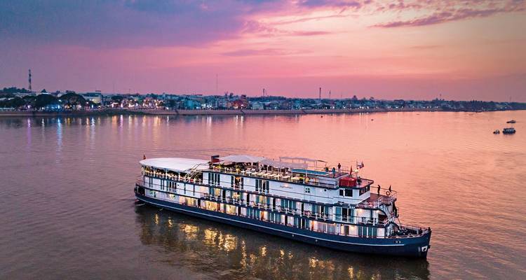 Kreuzfahrtschiff fährt bei Sonnenuntergang auf einem Fluss mit Stadtblick.