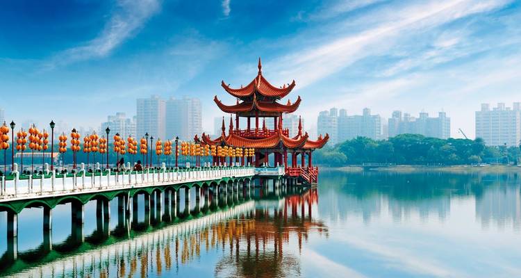 Ein traditioneller chinesischer Pavillon am Ufer eines Sees mit Stadtgebäuden im Hintergrund.