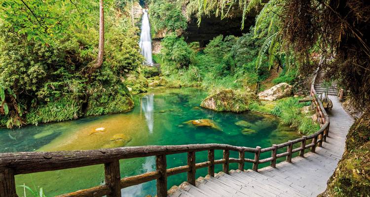 Ein malerischer Wasserfall, der in ein Becken stürzt, umgeben von üppigem Grün und einem Holzweg.