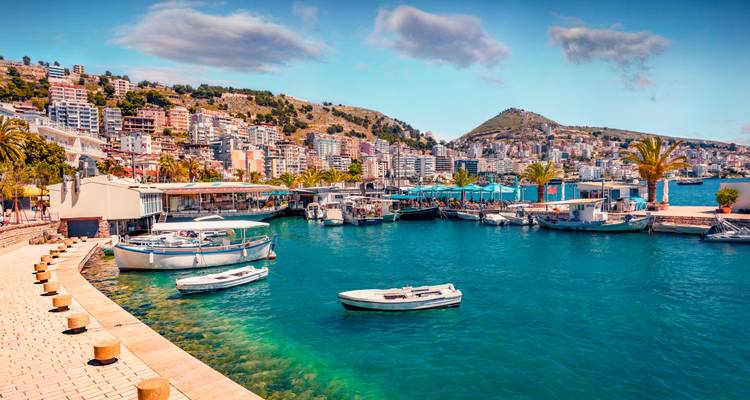 Paseo marítimo vibrante y puerto deportivo en Sarandá con aguas turquesas y el pueblo en la ladera como telón de fondo.