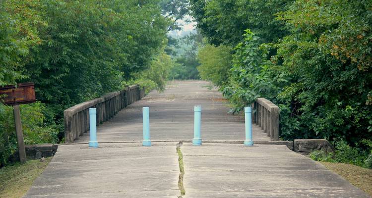 Betonbrücke blockiert durch blaue Pfosten, umgeben von dichtem grünem Laub.