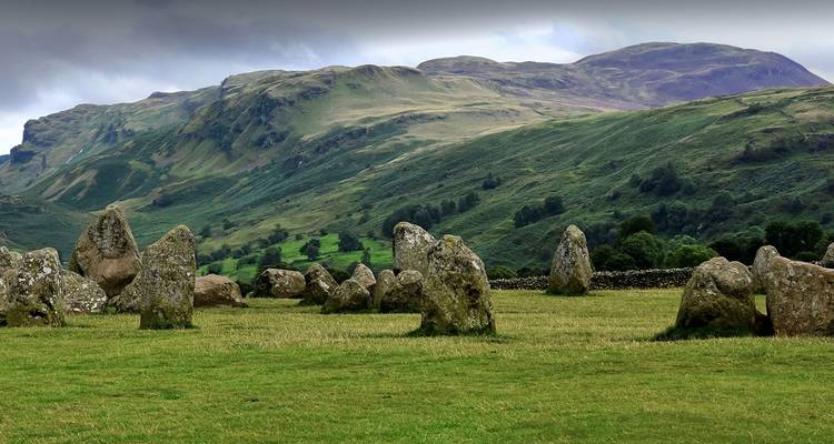 Ancient stone circle set in a wide grassy field with rolling Lake District hills in the background under an overcast sky.