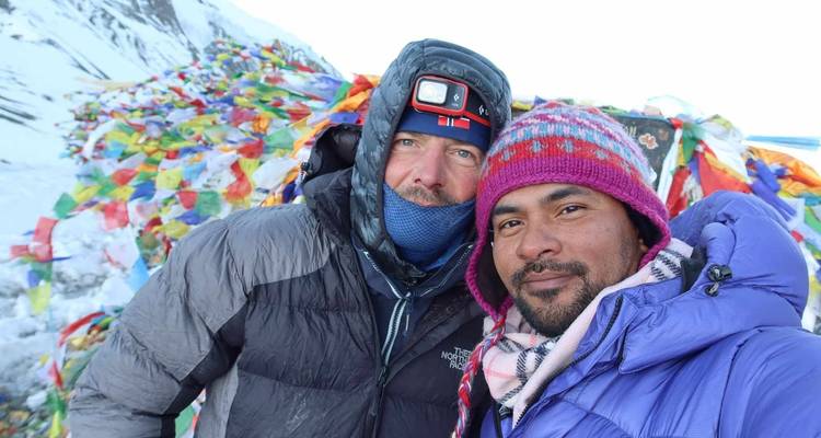 Zwei Männer in Winterkleidung mit Gebetsfahnen im Hintergrund.